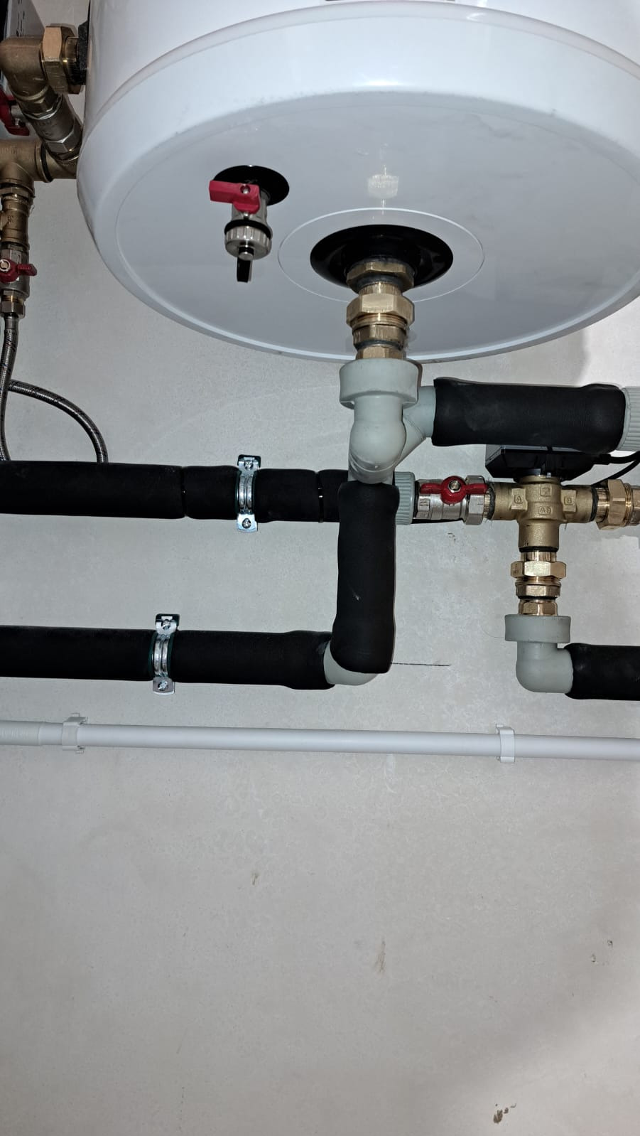 Biały zasobnik ciepłej wody użytkowej z widocznymi rurami doprowadzającymi i odprowadzającymi wodę, zaizolowanymi czarną otuliną, zamocowanymi do ściany za pomocą metalowych obejm.