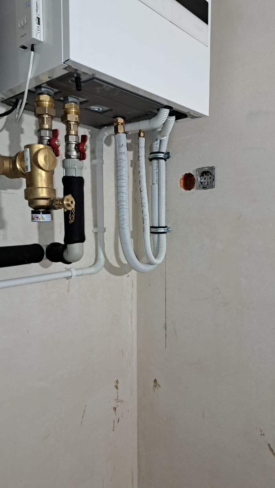 Nowy, biały piec gazowy zamontowany na ścianie, widoczne miedziane rury z zaworami, białe rury z izolacją, surowa ściana w tle przed wykończeniem.