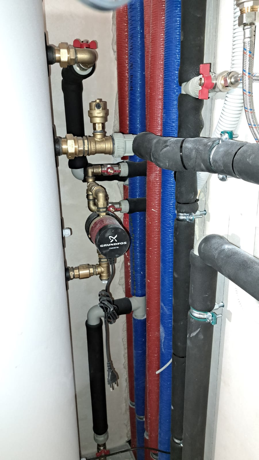 Instalacja hydrauliczna z pompą obiegową Grundfos, rurami w izolacji czarnej, oraz rurami czerwonymi i niebieskimi, widoczne zawory i złączki mosiężne.