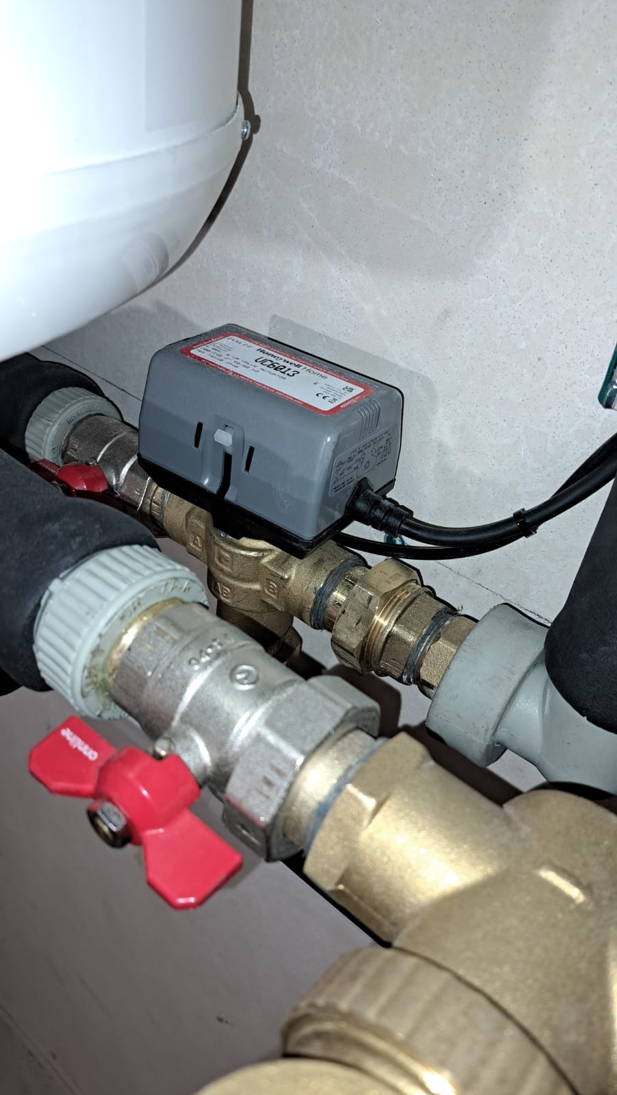 Szczegółowe ujęcie instalacji hydraulicznej z zaworami, rurami miedzianymi i szarym siłownikiem Honeywell UC568L3, zamontowanego na rurze, z widocznymi śrubami i złączkami.