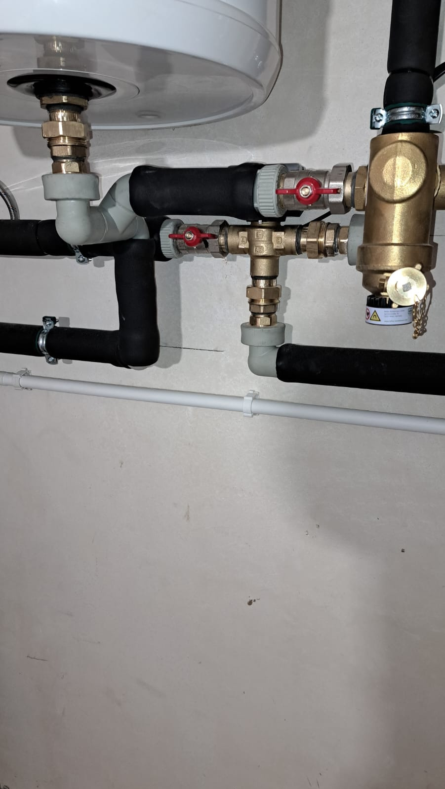 Fragment instalacji hydraulicznej z zaworami, rurami z izolacją i elementami mocującymi, widoczne przyłącze do zbiornika, szara ściana w tle.