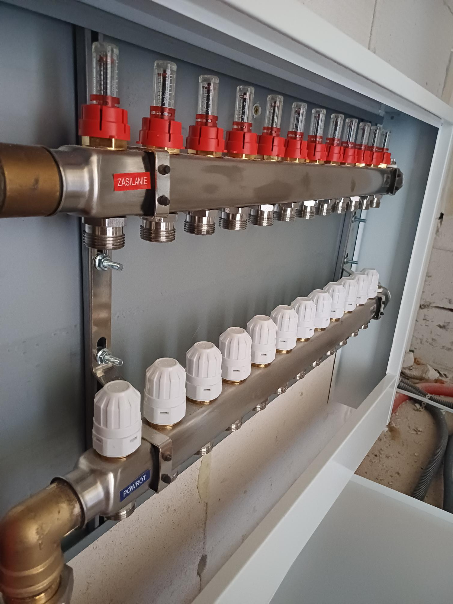 Rozdzielacz ogrzewania podłogowego z przepływomierzami i zaworami termostatycznymi, zamontowany w szafce instalacyjnej, widoczne napisy 'ZASILANIE' i 'POWRÓT'.