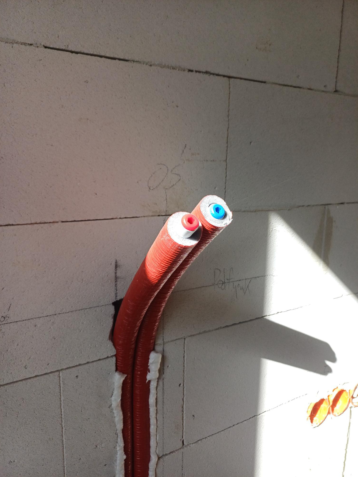 Dwie czerwone rury w izolacji, wystające ze ściany z betonu komórkowego, uszczelnione pianką montażową; widoczne końcówki rur z kolorowymi zaślepkami (czerwona i niebieska); cień na ścianie.