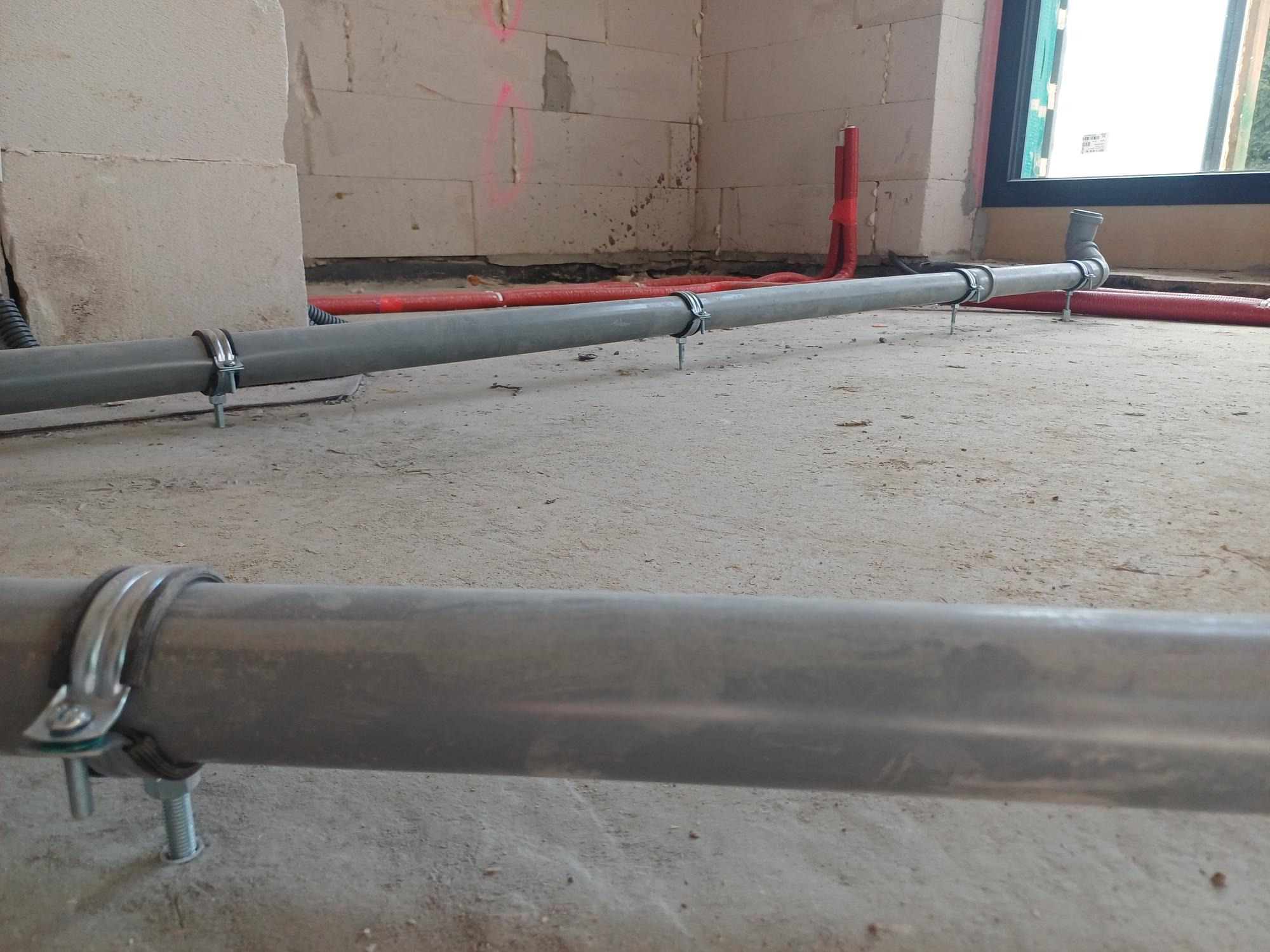 Szare rury kanalizacyjne, mocowane metalowymi uchwytami do surowej betonowej podłogi w trakcie budowy, widoczne czerwone rury grzewcze w tle.