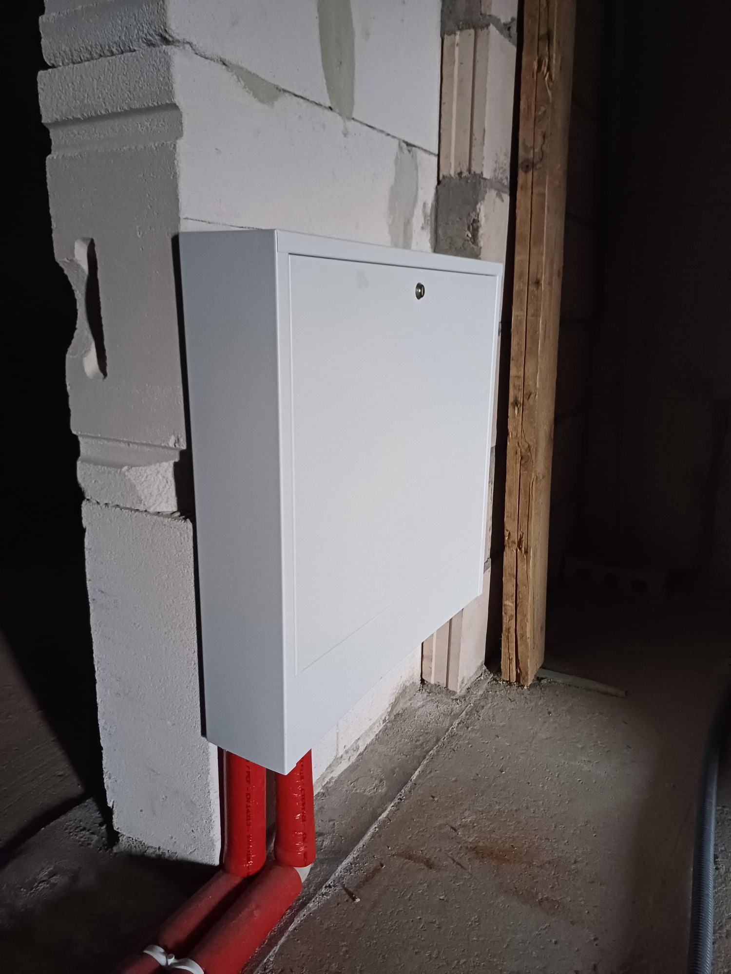 Biała skrzynka rozdzielcza z zabezpieczonym zamkiem, zamontowana na ścianie z betonu komórkowego, z widocznymi czerwonymi rurami instalacji grzewczej wchodzącymi do niej od dołu, w surowym stanie...