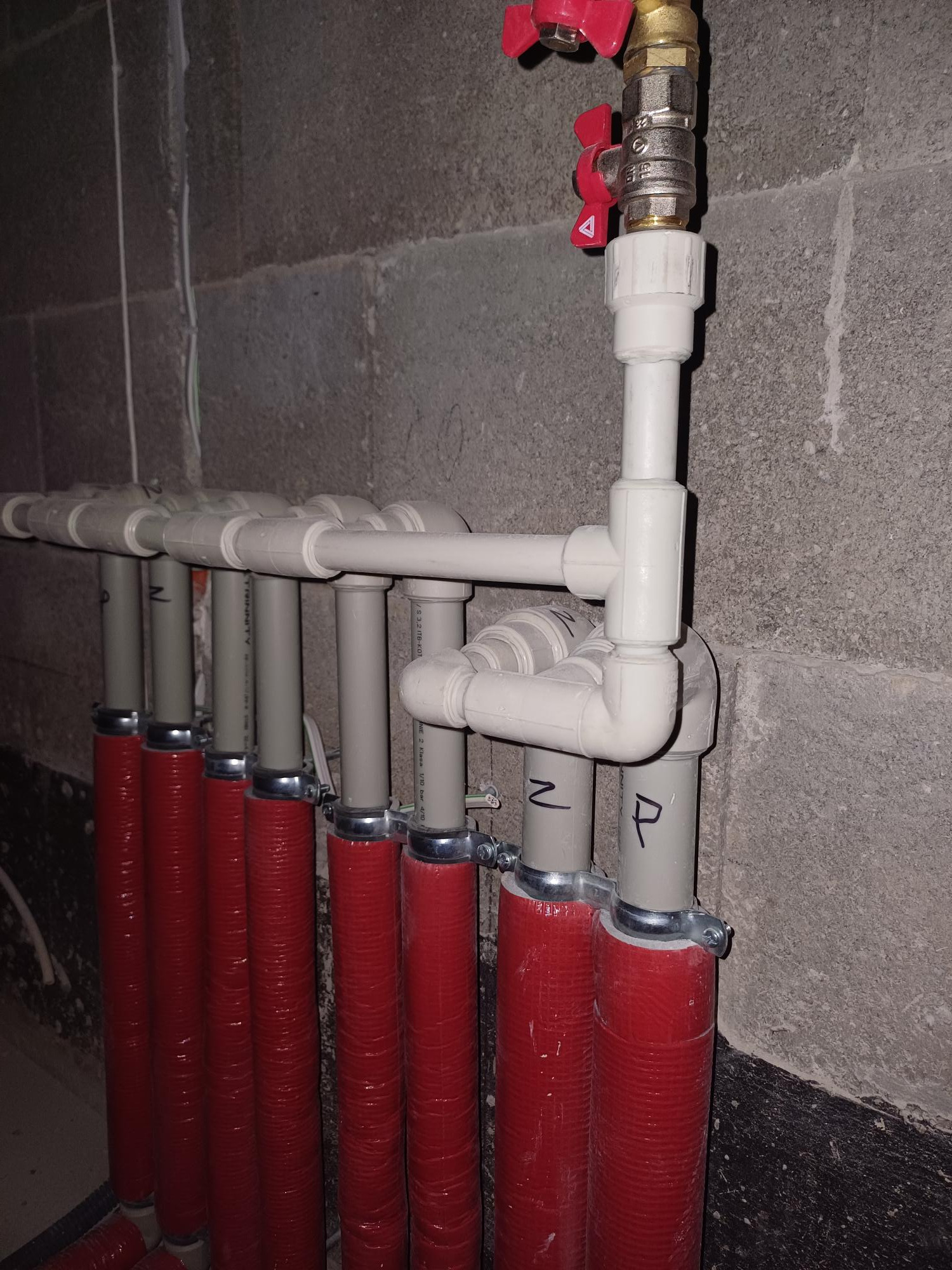 Szereg pionowych rur grzewczych w czerwonej izolacji, przymocowanych do surowej ściany z betonu komórkowego, z widocznymi oznaczeniami literowymi na szarych odcinkach rur i zaworem odcinającym...