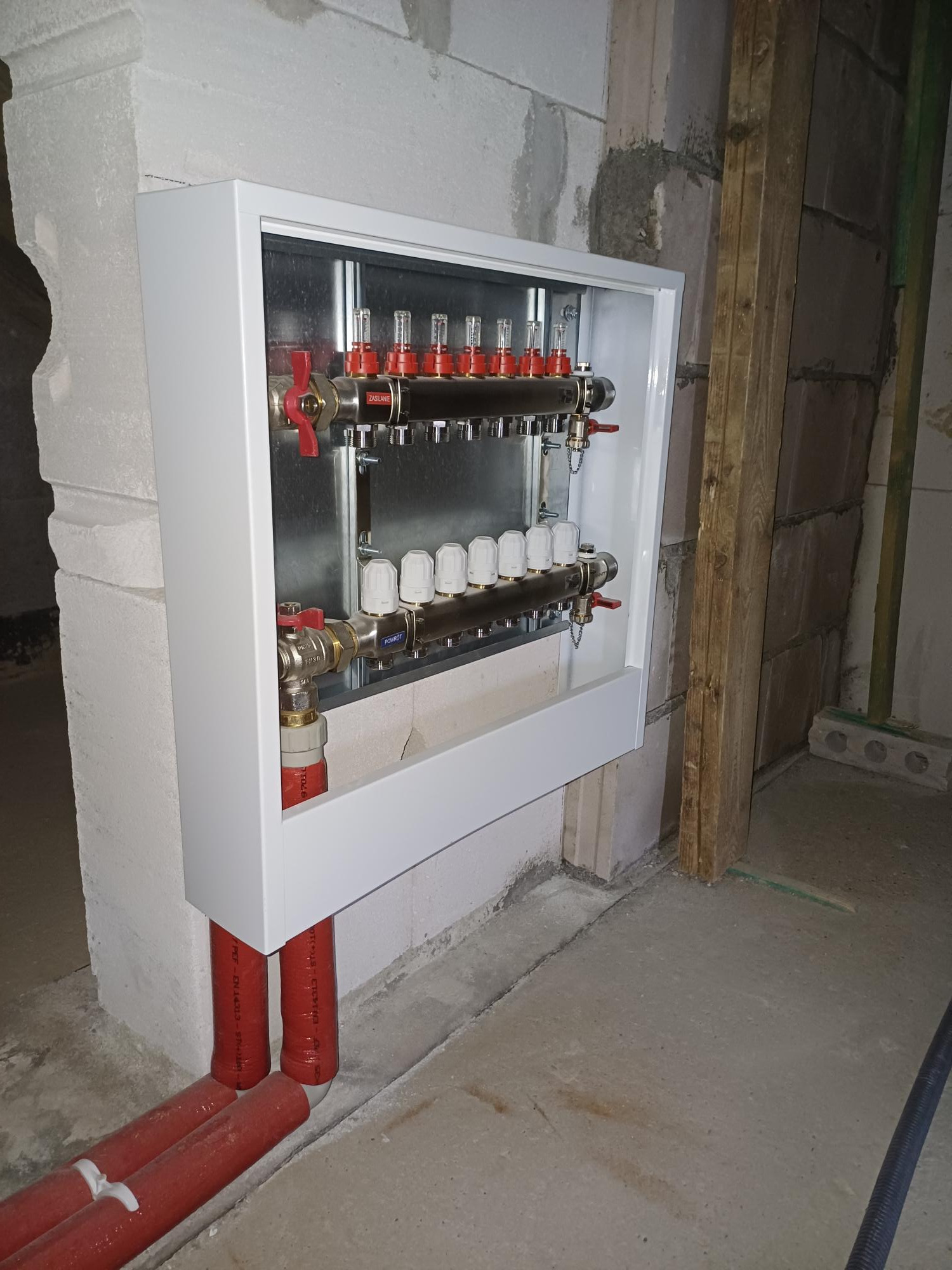 Rozdzielacz ogrzewania podłogowego w białej obudowie, z czerwonymi zaworami i rurami, zamontowany na surowej ścianie z betonu komórkowego w trakcie prac wykończeniowych.