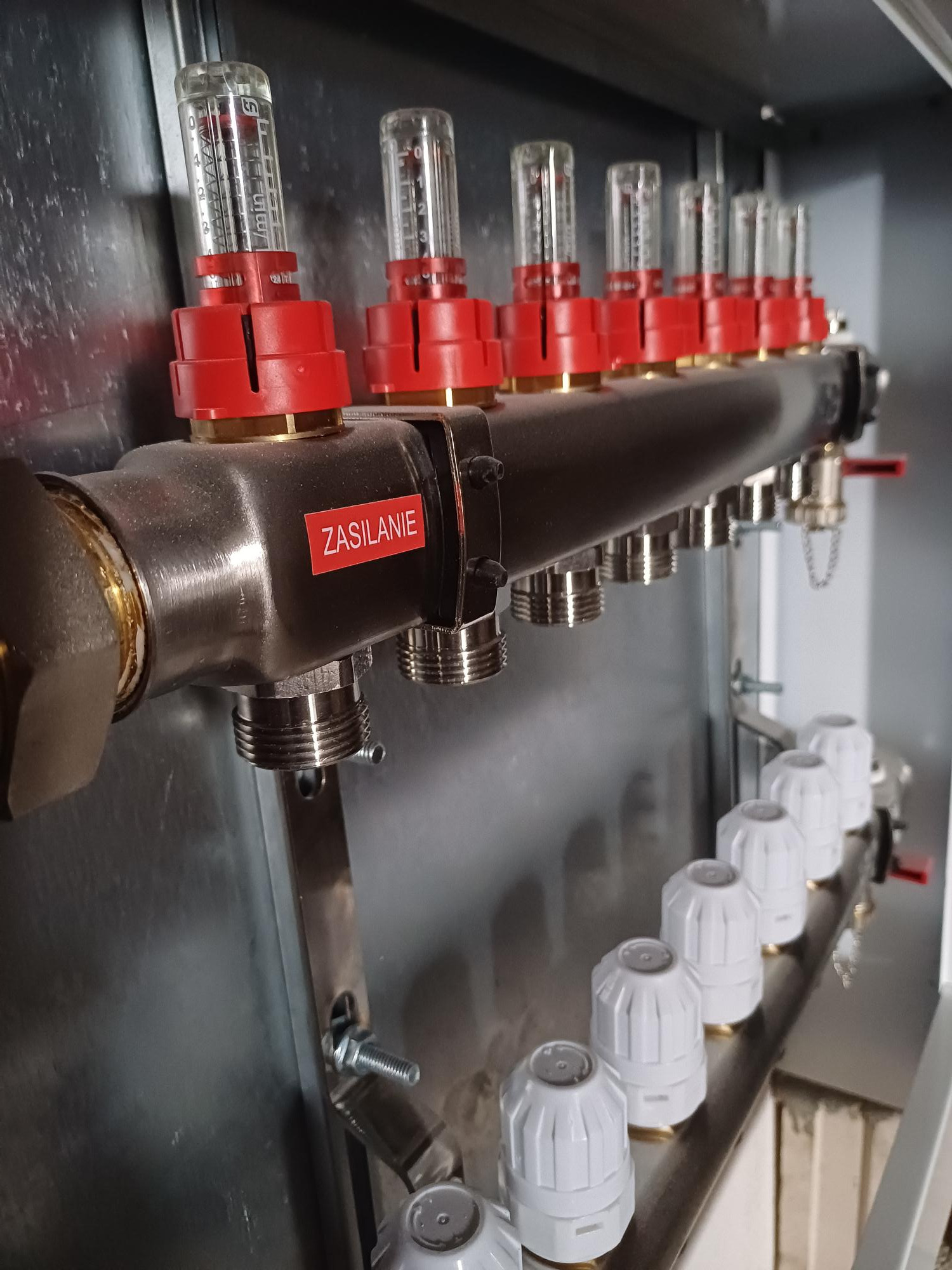 Rozdzielacz instalacji grzewczej z przezroczystymi przepływomierzami z czerwonymi elementami, zamontowany na metalowej płycie, z widoczną naklejką 'Zasilanie'.