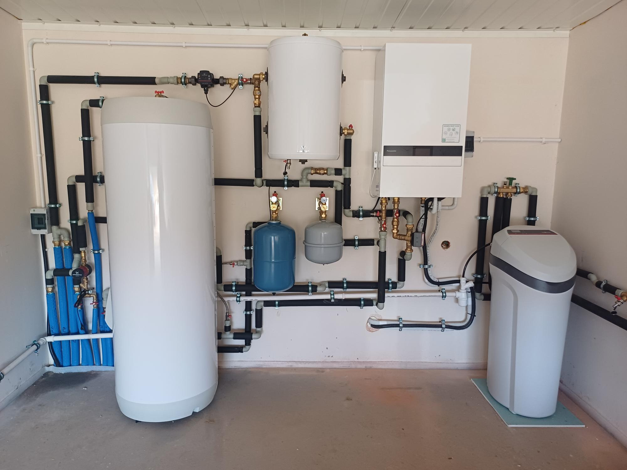 Kompletna instalacja grzewcza w kotłowni: dwa białe zbiorniki akumulacyjne, szaro-błękitne naczynia przeponowe, piec gazowy kondensacyjny Panasonic oraz kompaktowy zmiękczacz wody na podkładce.