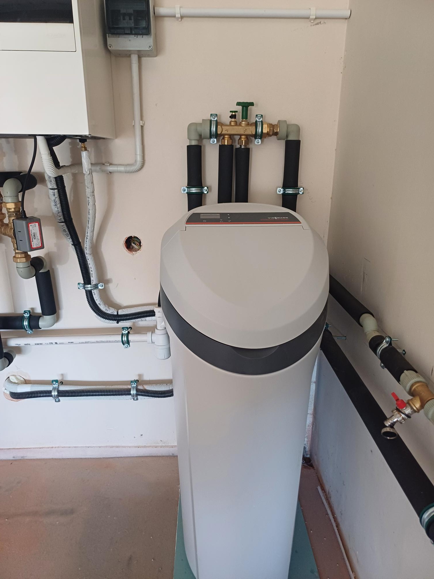 Centralny zmiękczacz wody Viessmann z podłączeniami hydraulicznymi, widoczne rury w izolacji, zawory i przewody elektryczne zamocowane do ściany za pomocą uchwytów.