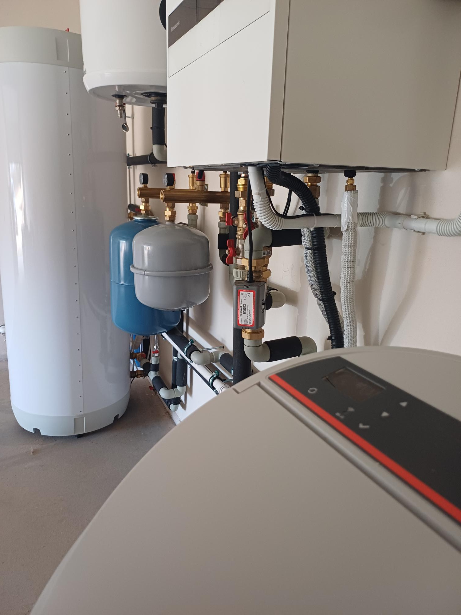 Kompleksowa instalacja: biały zbiornik, niebieski zbiornik wyrównawczy i panel sterowania z czerwonym akcentem, widoczne liczne rury i zawory.