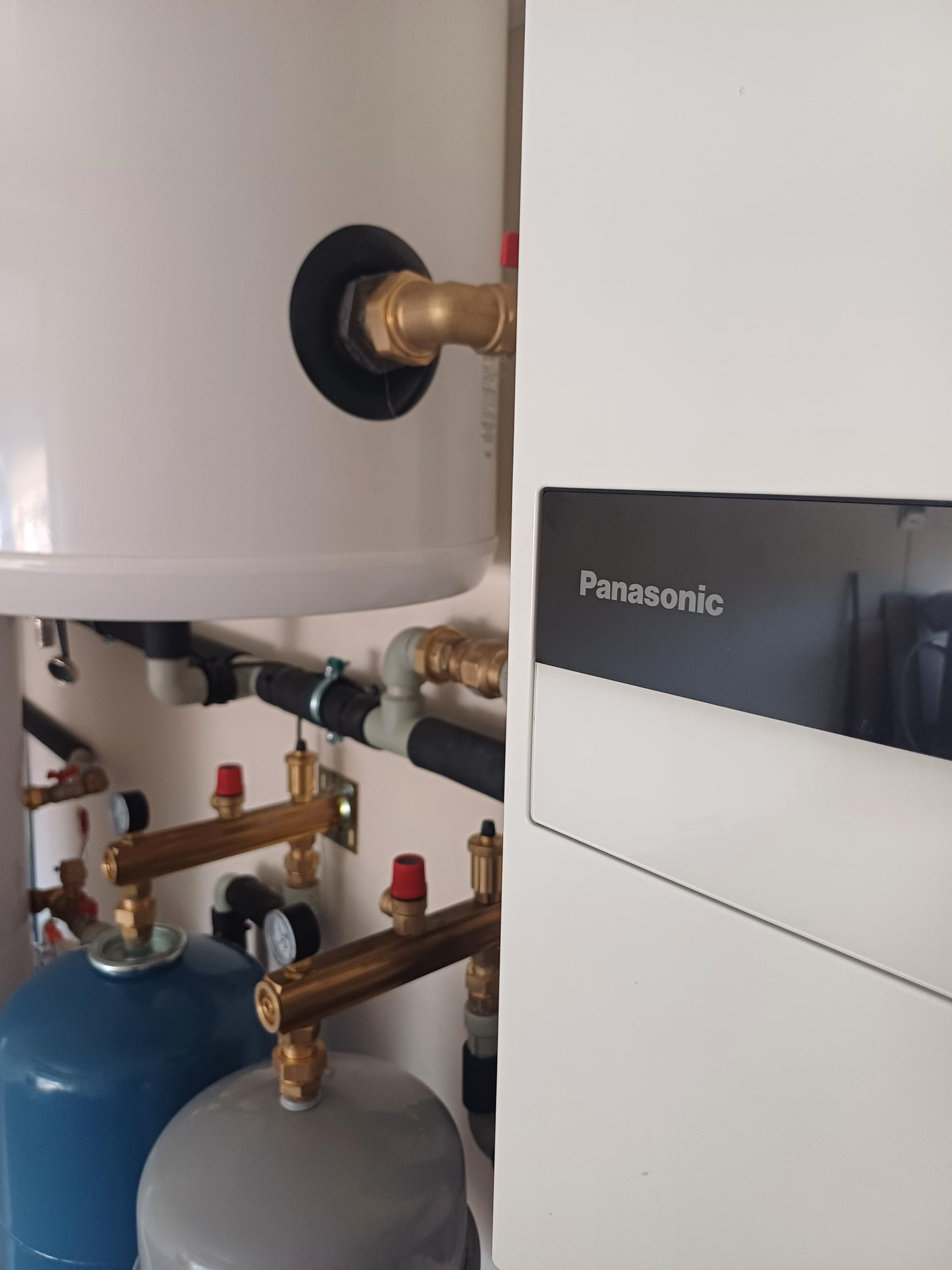 Biała jednostka wewnętrzna z logo Panasonic, widoczne przyłącza hydrauliczne z manometrami i zbiornikami wyrównawczymi w kolorze niebieskim i szarym.