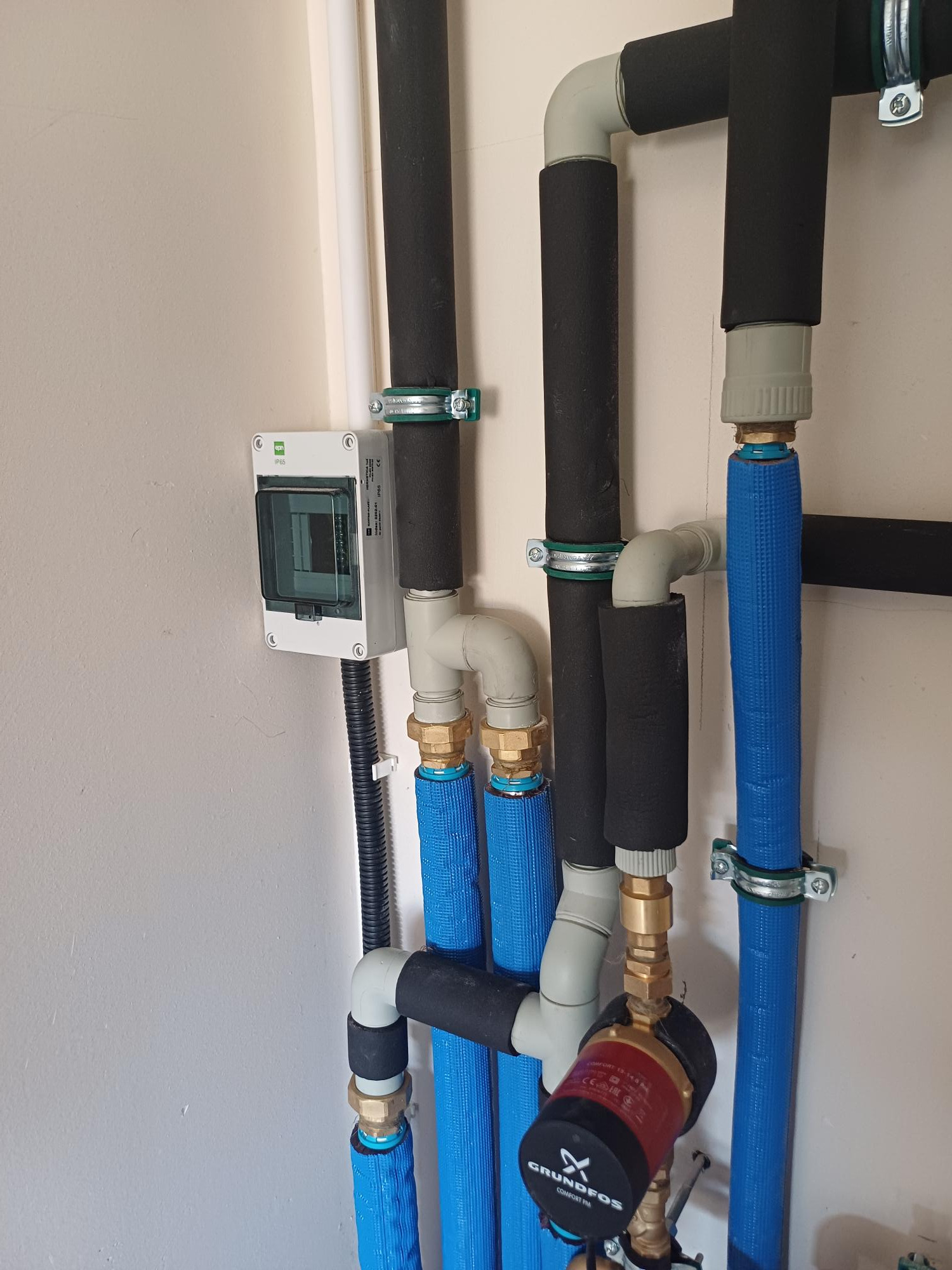 Szczegółowe ujęcie instalacji grzewczej z rurami w niebieskiej izolacji, czarnymi rurami z otuliną i pompą obiegową Grundfos Comfort PM, zamontowanej na jasnej ścianie obok skrzynki elektrycznej.
