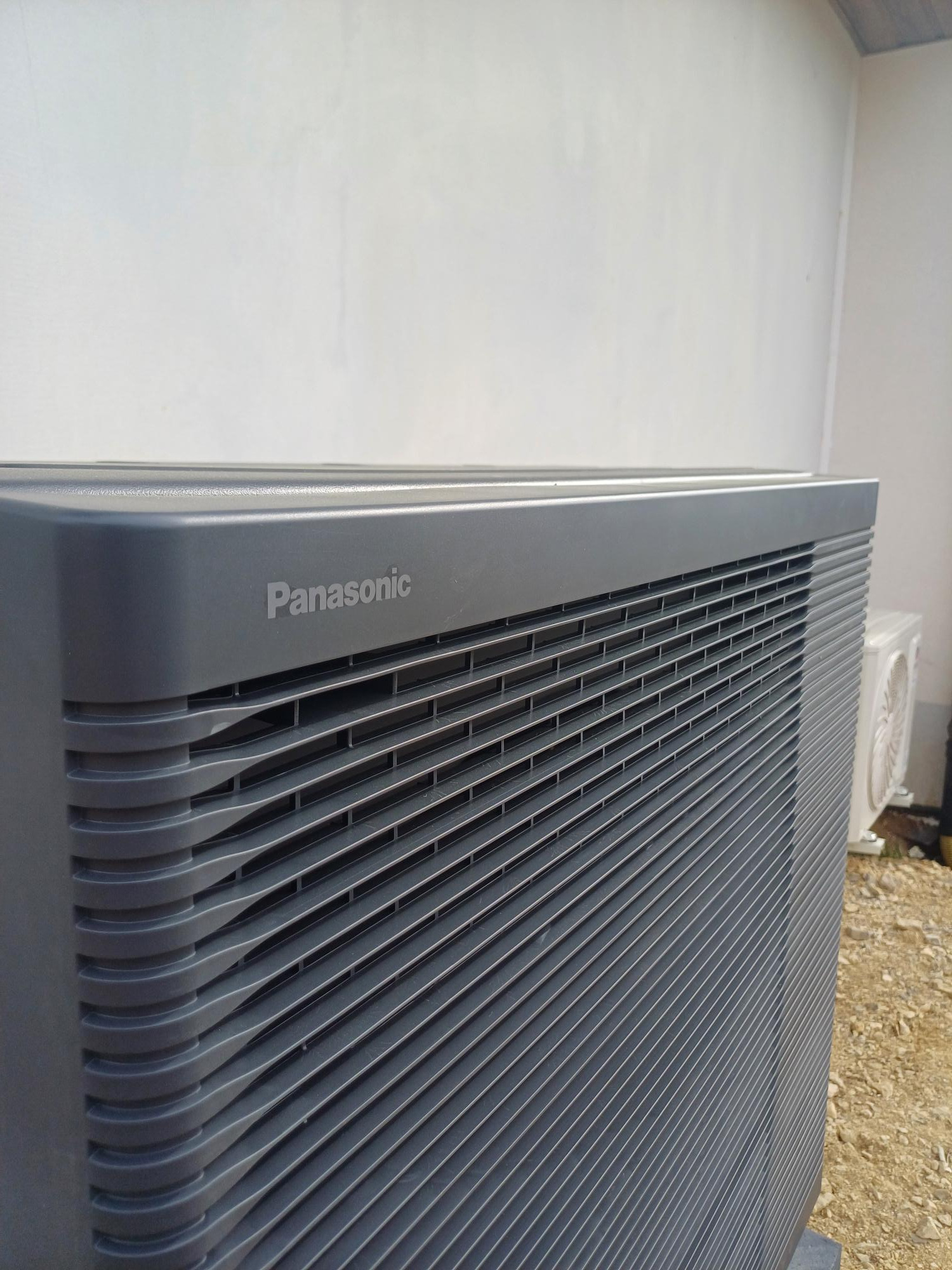 Szara jednostka zewnętrzna pompy ciepła Panasonic z widocznymi lamelami wentylacyjnymi i logo, na tle białej ściany i żwirowego podłoża, z drugą jednostką w tle.