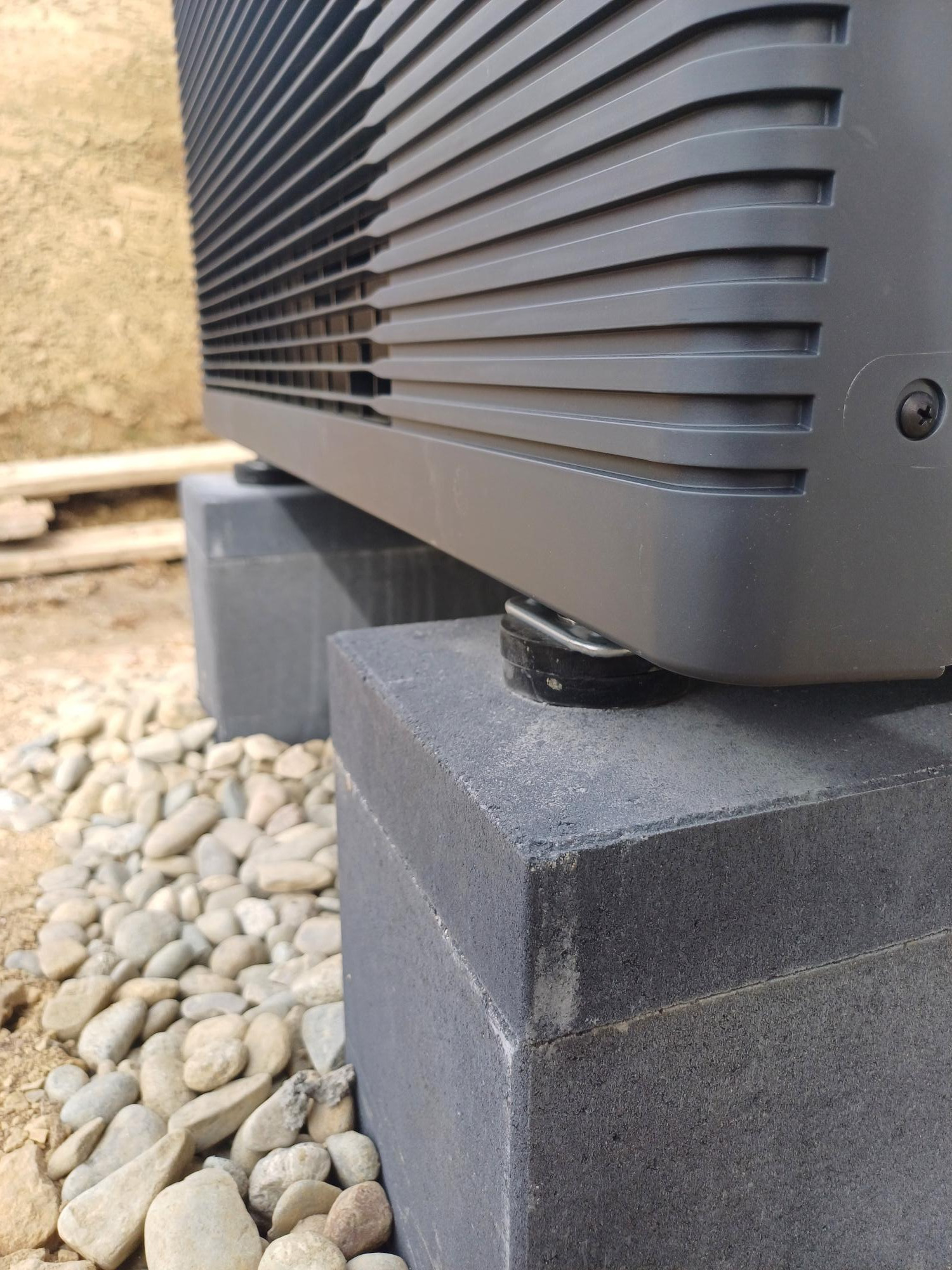 Szara jednostka zewnętrzna urządzenia grzewczego, prawdopodobnie pompy ciepła, ustawiona na dwóch betonowych bloczkach na żwirowym podłożu. Widoczny fragment elewacji budynku w tle.