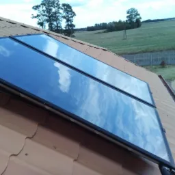 Dwa panele słoneczne zamontowane na brązowym dachu z dachówki ceramicznej, odbijające błękitne niebo z chmurami, w tle zielone pola i drzewa.