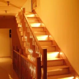 Jasne schody z drewna z podświetleniem LED na każdym stopniu. Metalowe słupki balustrady. Ciepłe oświetlenie wnętrza. Widok z dołu na górę.