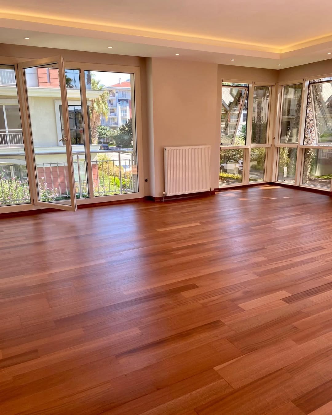 Wypolerowana, parkietowa podłoga z egzotycznego drewna w przestronnym, jasnym salonie z dużymi oknami balkonowymi i widokiem na zieleń miejską.