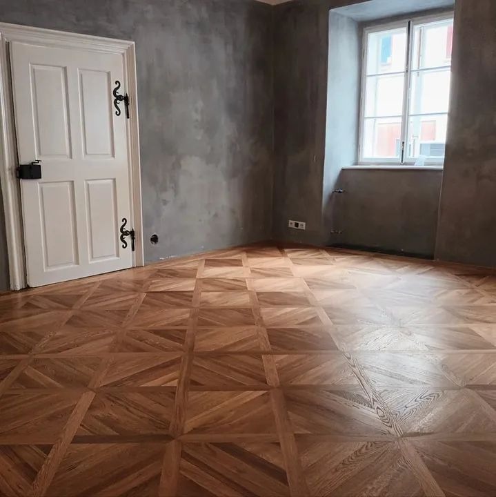 Parkiet w geometryczny wzór w wykończonym pokoju z szarą ścianą i białymi drzwiami z ozdobnymi zawiasami.