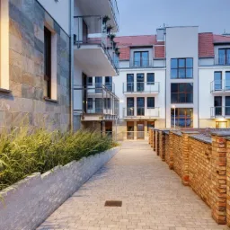 Nowoczesny budynek wielorodzinny z balkonami i murem z cegły, oświetlony wieczornym światłem. Widok na elewację z kamienia i zieleni wzdłuż ścieżki z kostki.