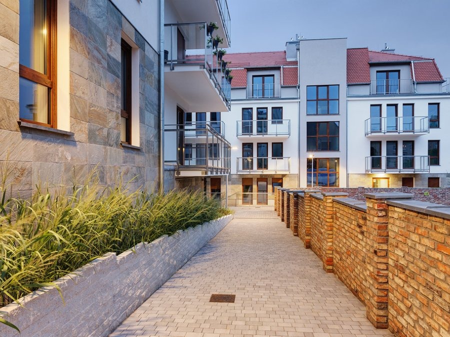 Nowoczesny budynek wielorodzinny z balkonami i murem z cegły, oświetlony wieczornym światłem. Widok na elewację z kamienia i zieleni wzdłuż ścieżki z kostki.