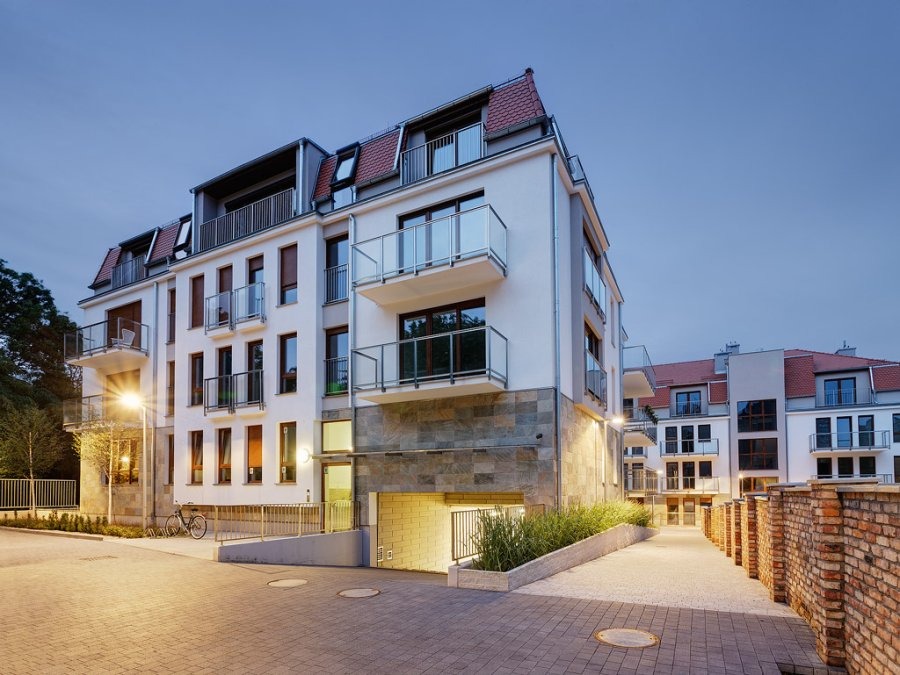 Nowoczesny budynek apartamentowy z balkonami i podziemnym garażem, oświetlony lampą uliczną, z chodnikiem wyłożonym kostką brukową i elementami zieleni.