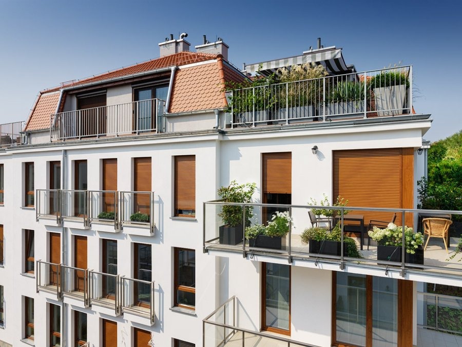 Biały budynek wielorodzinny z balkonami i roletami zewnętrznymi, rośliny w donicach na balkonach i tarasie na dachu. Widok z dołu na elewację.