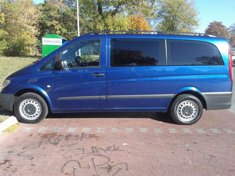 Niebieski bus Mercedes Vito z relingami dachowymi, zaparkowany na chodniku z kostki. Widok z boku, w tle zieleń drzew i pojemnik na odzież.