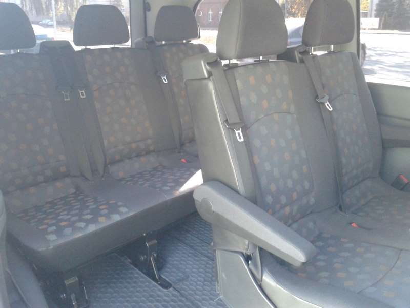 Wnętrze busa z trzema rzędami siedzeń w kolorze szarym z delikatnym wzorem. Widoczne zagłówki i pasy bezpieczeństwa. Czyste i zadbane wnętrze pojazdu.