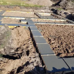 Zalane betonem szalunki fundamentowe na piaszczystym gruncie, widoczne wystające pręty zbrojeniowe.