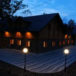 Oświetlony budynek z drewnianą elewacją i kamienną podmurówką, z brukowanym chodnikiem w geometryczny wzór i lampami kulami, widziany o zmierzchu.