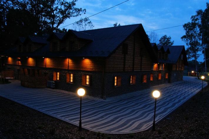 Oświetlony budynek z drewnianą elewacją i kamienną podmurówką, z brukowanym chodnikiem w geometryczny wzór i lampami kulami, widziany o zmierzchu.