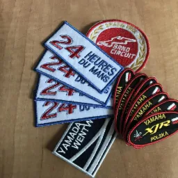 Kolekcja naszywek z motywami wyścigowymi i motocyklowymi, w tym '24 Heures du Mans', 'Brno Circuit' i 'Yamaha XJR Polska', ułożonych na drewnianym tle.