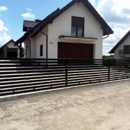 Nowoczesna, pozioma brama wjazdowa w kolorze czarnym, z widocznym mechanizmem przesuwnym, na tle domu z brązową stolarką okienną i numerem 25A.