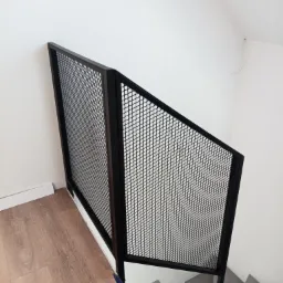 Minimalistyczna, czarna, metalowa balustrada schodowa z wypełnieniem z siatki, zamontowana w nowoczesnym wnętrzu z szarymi schodami i panelami podłogowymi.