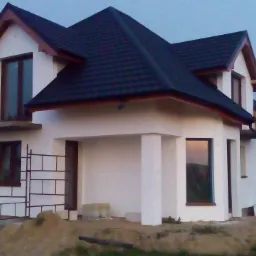 Nowy dom z ciemną dach&oacute;wką i białymi ścianami, widoczne rusztowania przy bocznej ścianie. Dom w stanie surowym zamkniętym, otoczony ziemią.