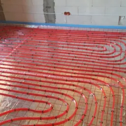 Instalacja ogrzewania podłogowego z czerwonych rur na metalowej siatce, widoczne przyłącza elektryczne w ścianie z bloczków.