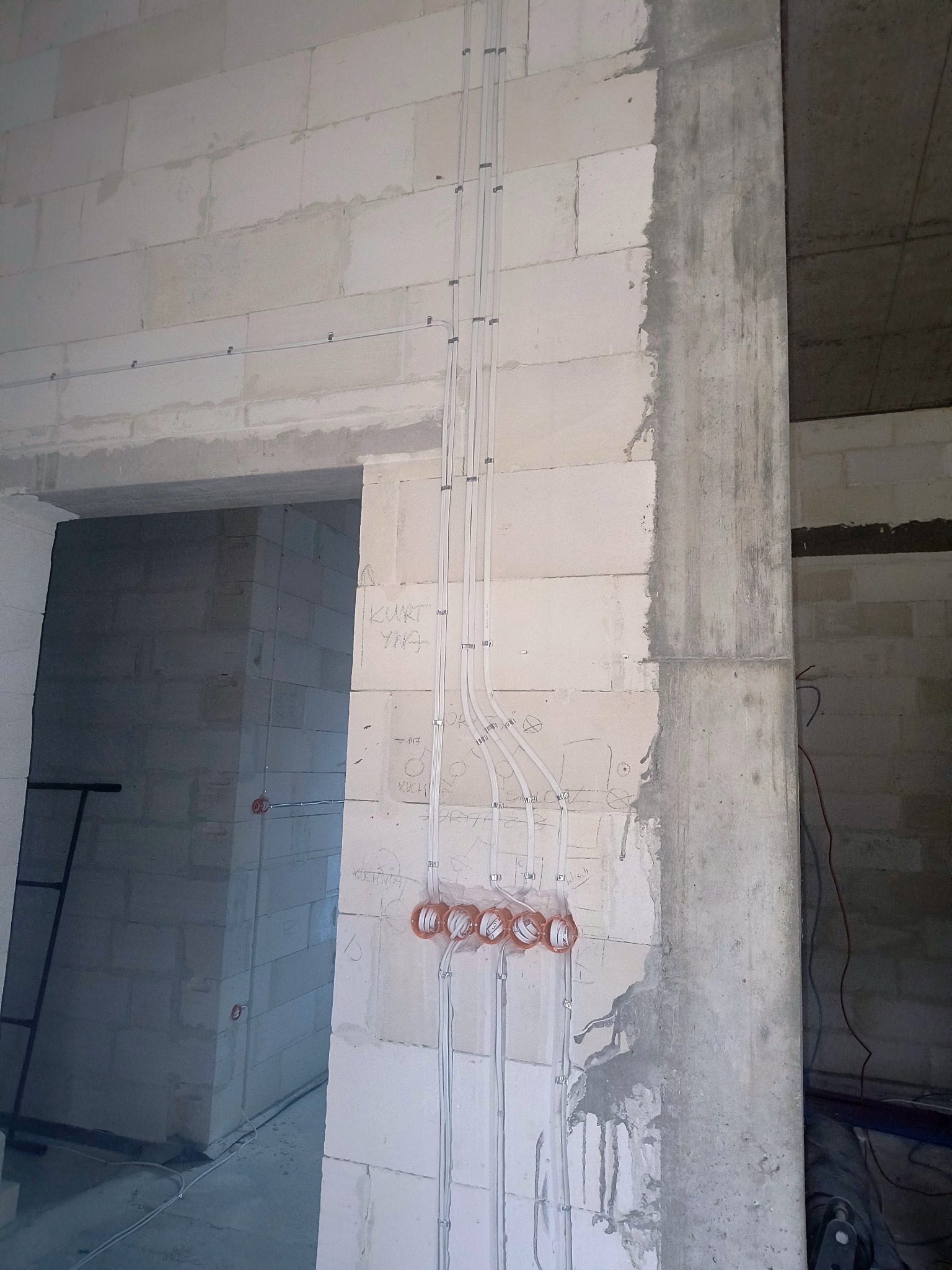 Surowa ściana z betonu komórkowego z poprowadzonymi przewodami elektrycznymi w peszlach i puszkami instalacyjnymi. Widoczne napisy na ścianie. Etap prac instalacyjnych.