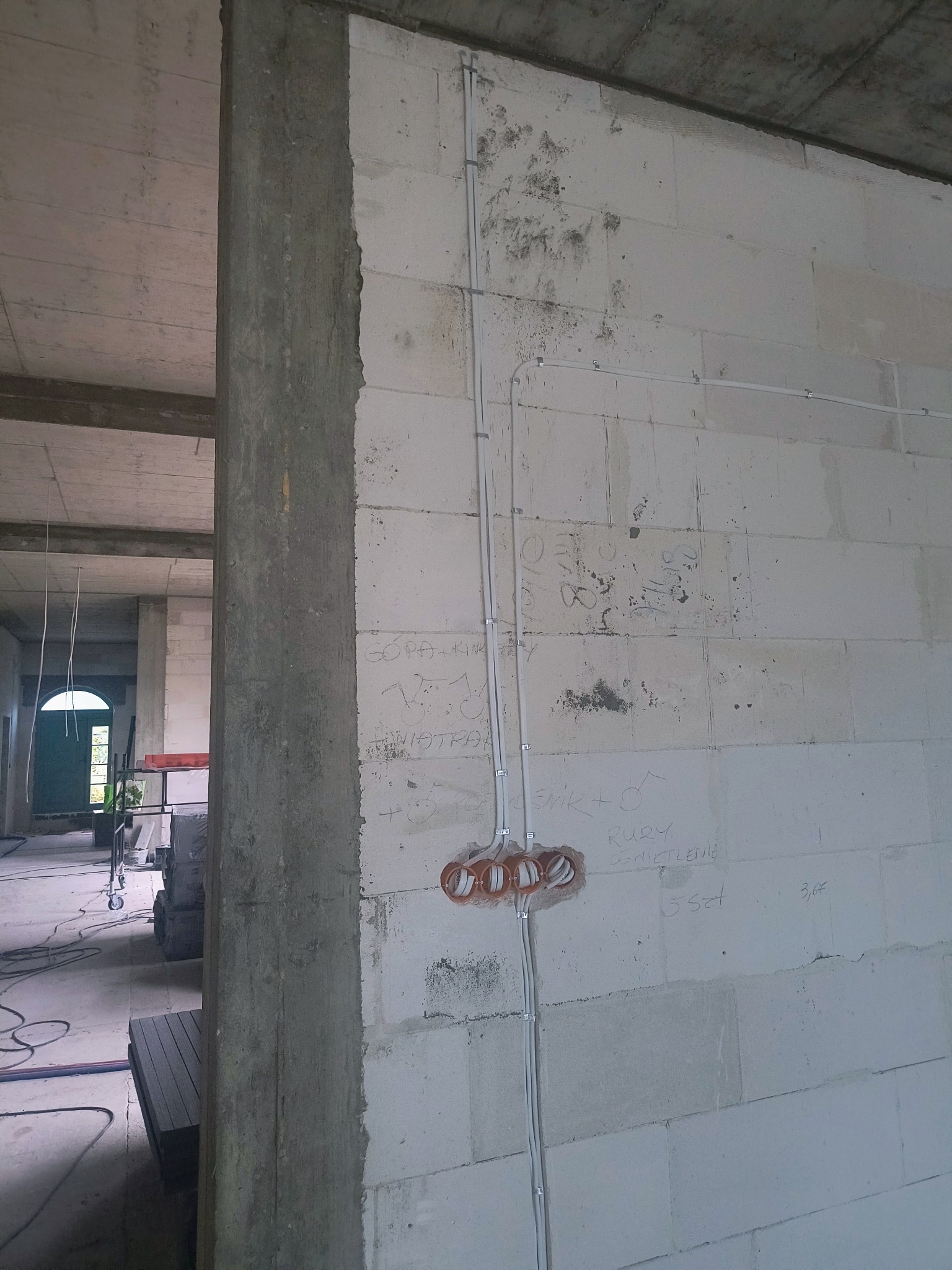 Surowa ściana z białego betonu komórkowego z widoczną instalacją elektryczną w postaci białych kabli i pomarańczowych puszek pod gniazdka. W tle widoczny surowy stan budynku.