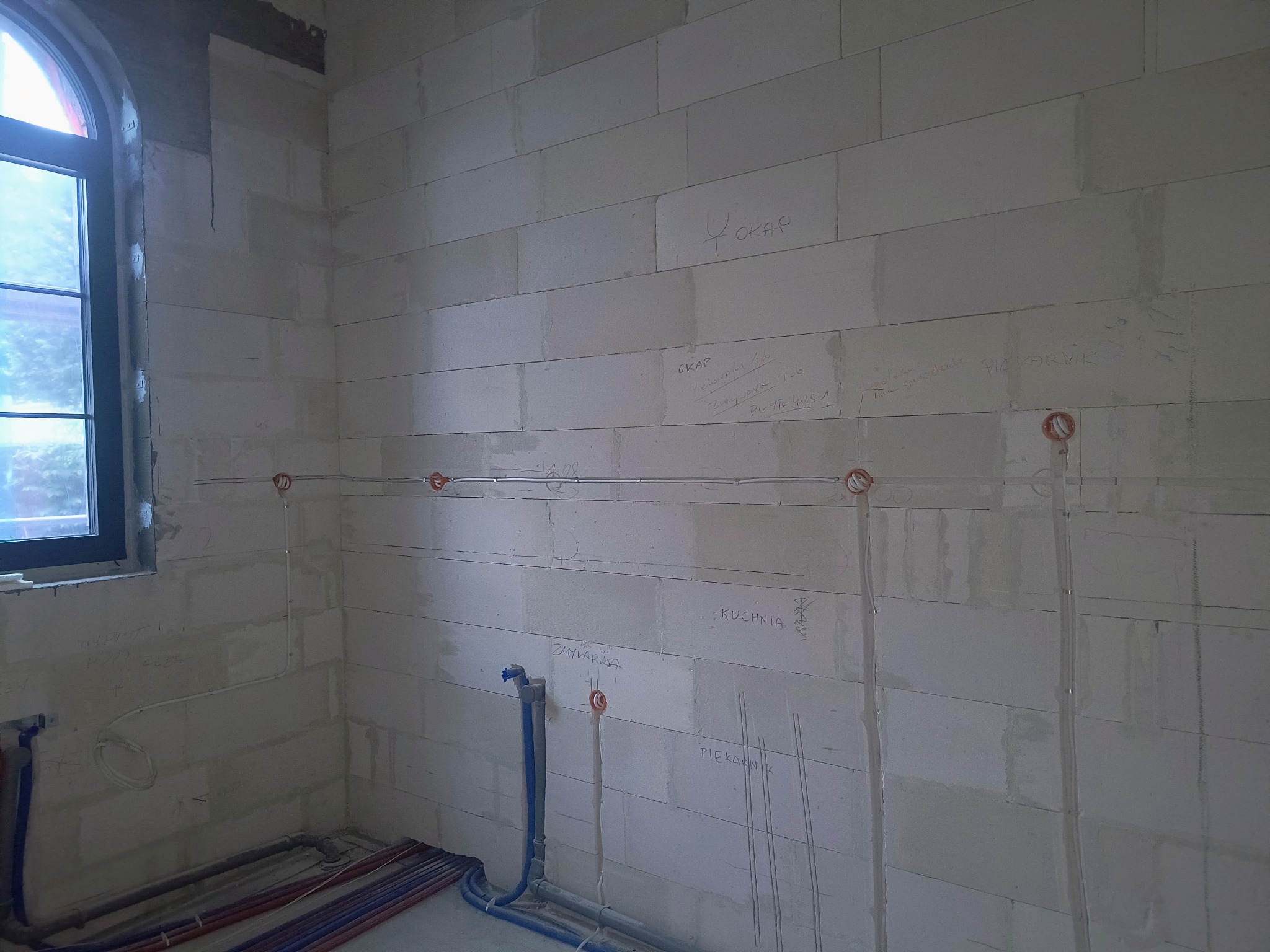 Wnętrze w trakcie prac instalacyjnych: widoczne przewody elektryczne i rury wodne na ścianach z betonu komórkowego z naniesionymi odręcznymi oznaczeniami.