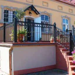 Czarna, metalowa balustrada z dekoracyjnymi okręgami, zamontowana na tarasie i schodach wejściowych do domu o żółtej elewacji, z widocznymi donicami z roślinami.