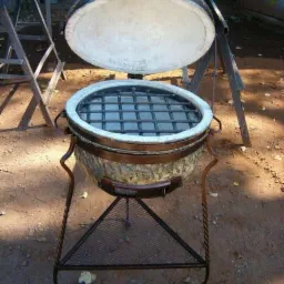 Ceramiczny grill ogrodowy typu Kamado z otwartą pokrywą, osadzony na metalowym, kutym stojaku z trójkątną półką z siatki, widoczne detale wykończenia i spawania.
