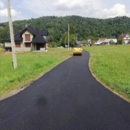 Walec drogowy ubija świeżo wylany asfalt na wiejskiej drodze, w tle domy i zalesione wzgórza.