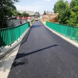 Świeżo położona, czarna nawierzchnia asfaltowa na moście z zielonymi barierkami, w tle budynki i stos drewna, w oddali walec drogowy.