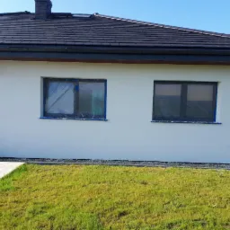 Biała elewacja domu jednorodzinnego z dwoma oknami zabezpieczonymi folią malarską, przygotowana do malowania, widok z poziomu trawnika.