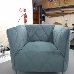 Turkusowy fotel z pikowanym wzorem rombów, stojący w warsztacie z gaśnicą na ścianie i maszyną do szycia w tle.