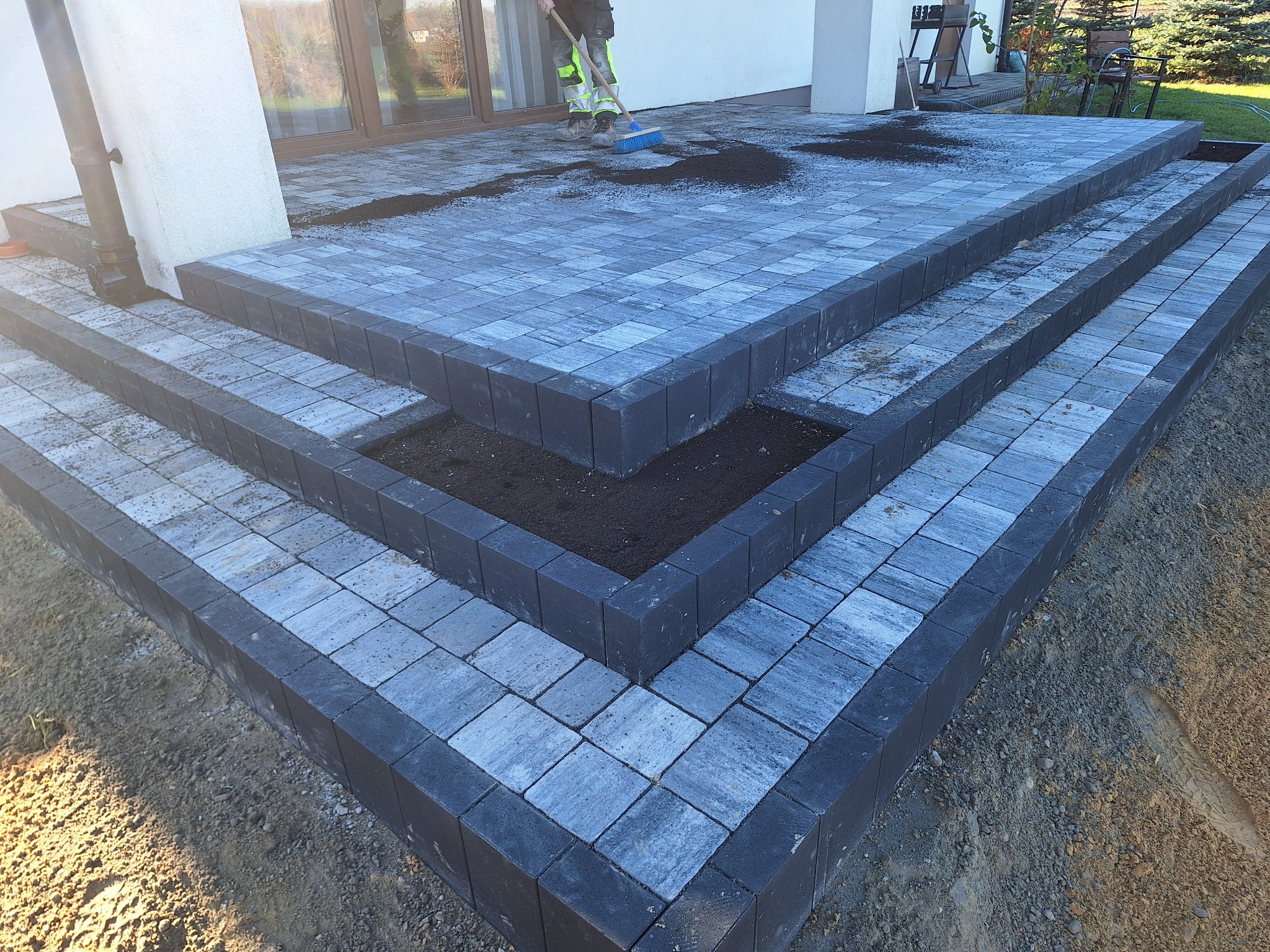 Schody z kostki brukowej w odcieniach szarości, z czarnymi obrzeżami, prowadzące do domu. Osoba zamiata powierzchnię. Widoczne przygotowane miejsca na rośliny.