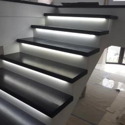 FHU Jarko - Nowoczesne schody z czarnymi stopniami podświetlonymi listwami LED, widok z dołu, jasne ściany i podłoga w tle.