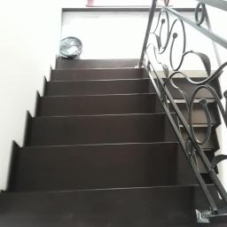 FHU Jarko - Ciemne schody z metalową balustradą o dekoracyjnym wzorze, widok z dołu, na górze schodów widoczny wentylator.