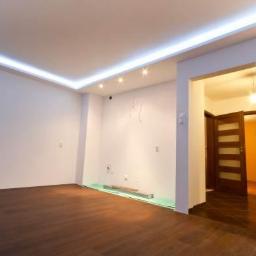 FHU Jarko - Jasne wnętrze po remoncie z drewnianą podłogą, listwami LED na suficie i widokiem na sąsiednie pomieszczenie przez otwarte drzwi.